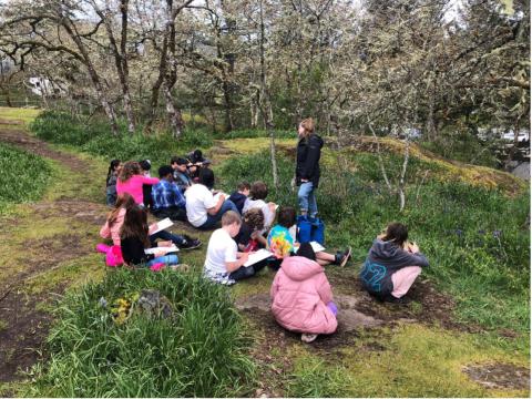 A group of students sitting on the ground outdoors, working on clipboards while a teacher stands and speaks to them.